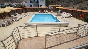 A swimming pool with sun loungers and umbrellas, surrounded by a stone wall and a view of a mountain. - Beautiful three bedroom apartment with shared swimming pool and WIFI (Puerto de Mogan)