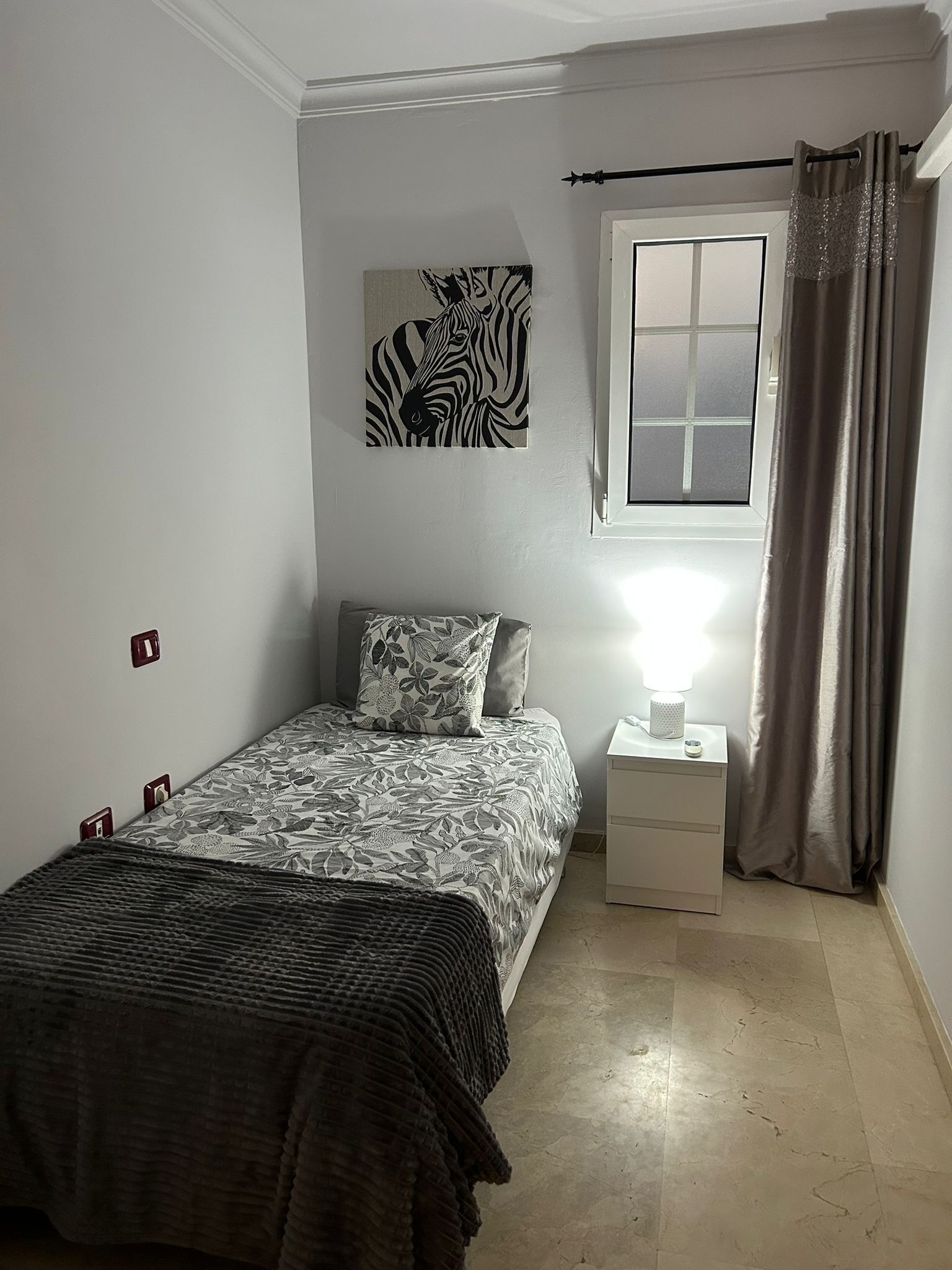 A bedroom with a bed, a nightstand, a lamp, a window with curtains, and a zebra artwork on the wall.