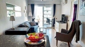 A modern living room with a sofa, a dining table with a bowl of fruit, a TV, and a ceiling fan. -  Beautiful Two bedroom apartment with spacious terrace, swimming pool and WIFI (Puerto de Mogan)