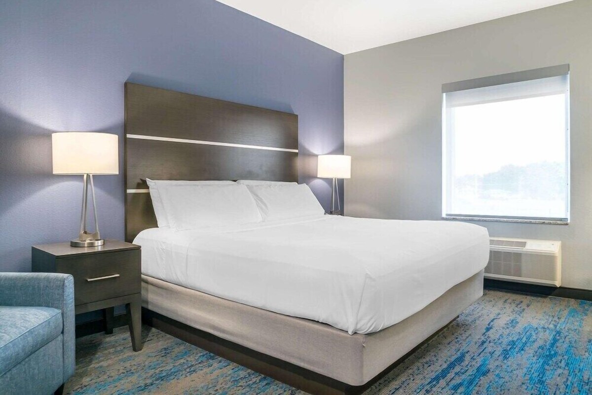 Habitación de hotel con cama, dos mesitas de noche con lámparas, una silla, un ventanal grande y piso alfombrado.