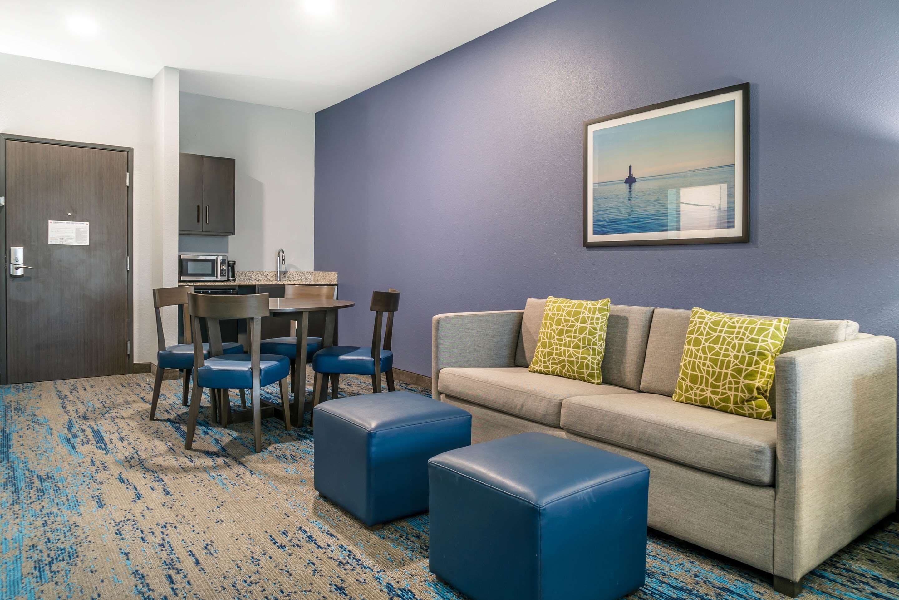 A hotel room with a gray sofa, blue ottomans, a dining table with chairs, a kitchen area, and a framed picture on the wall.