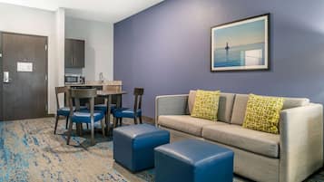 A hotel room with a gray sofa, blue ottomans, a dining table with chairs, a kitchen area, and a framed picture on the wall.