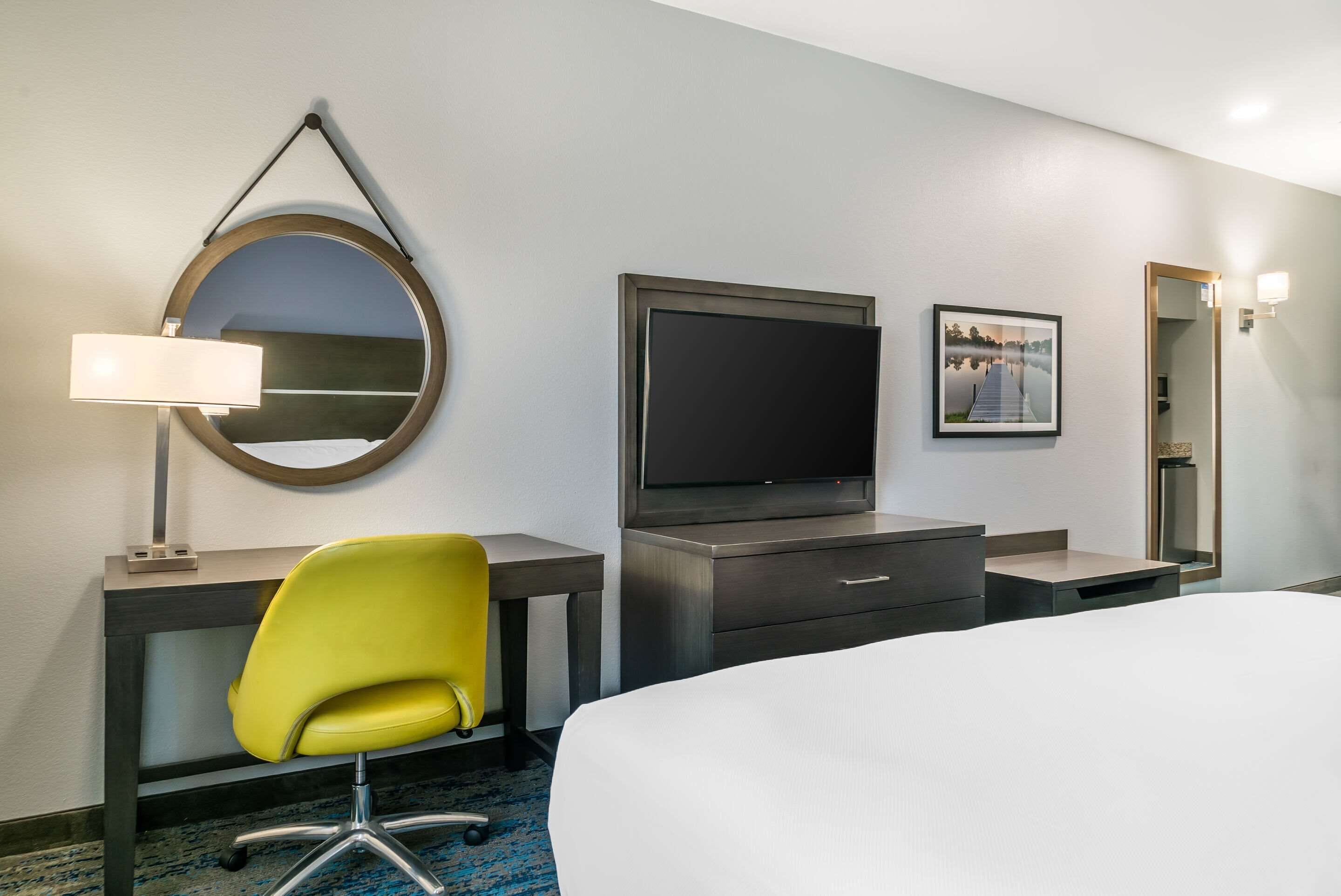 A hotel room with a bed, a desk with a chair, a television, a mirror, and a framed picture on the wall.