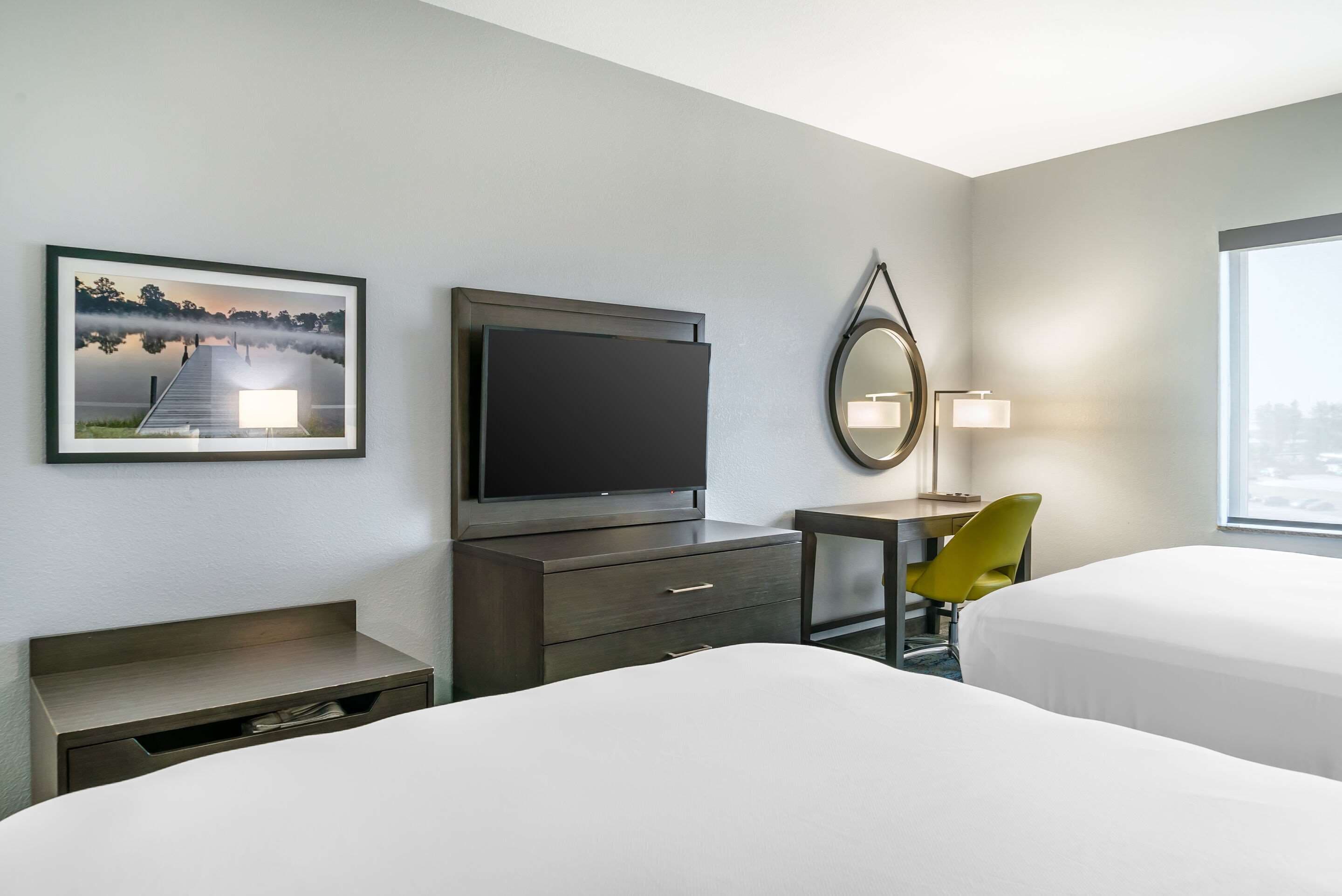 A hotel room with two beds, a flat-screen TV, a desk with a chair, a lamp, and a framed picture on the wall.