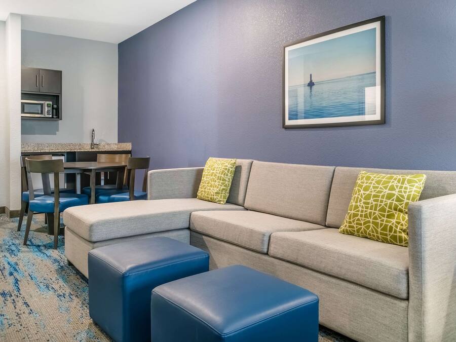 A modern living room with a sectional sofa, two blue ottomans, a dining area with a table and chairs, and a kitchen with a microwave.