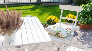 Un juego de mesa y sillas blancas en un patio empedrado, con un césped y flores amarillas al fondo.