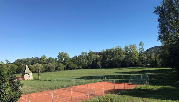 A tennis court with a red surface, surrounded by a green field and trees.