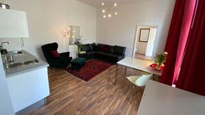 A modern living room with a black sofa, a green armchair, and a small table. There's a kitchen area with a sink and stove. The room has wooden flooring and a red curtain. - Excellently located 2-room apartment with balcony and high-speed internet in Mitte (Berlin)