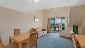 A hotel room with a sofa, a dining table, chairs, a large window with a view, and a clock on the wall.