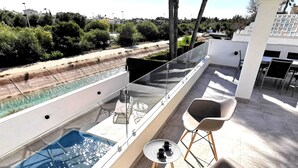 Une terrasse sur le toit dotée d’une balustrade en verre, une piscine, deux chaises et une petite table sur laquelle se trouvent une bouteille et une tasse.