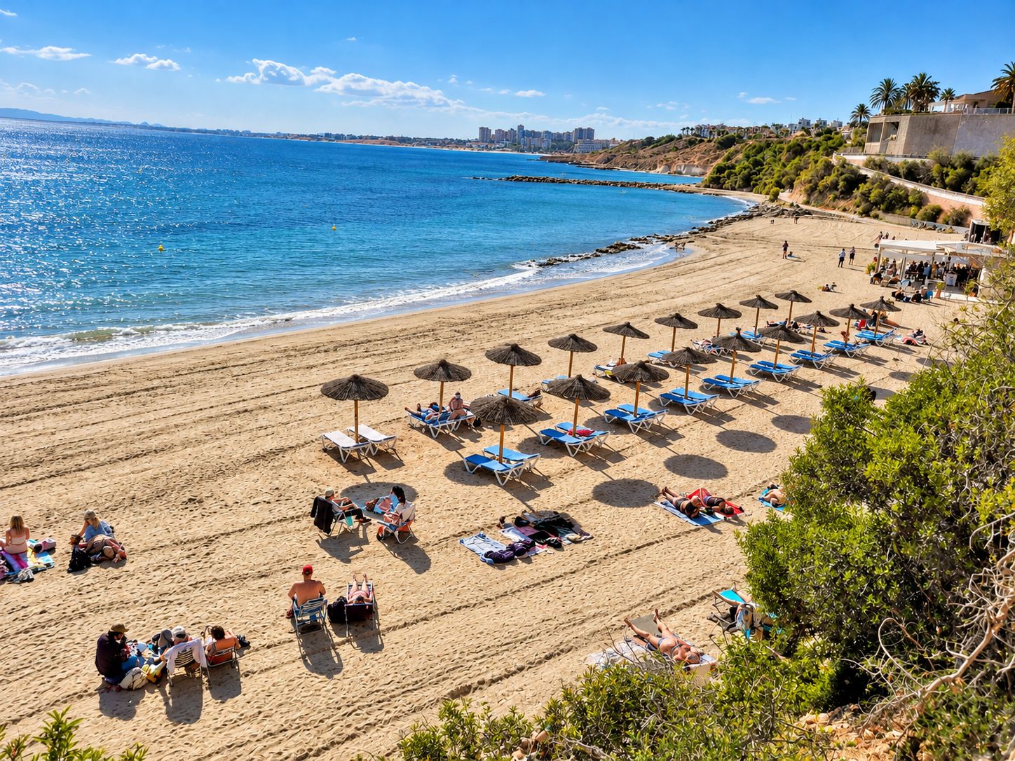 A beach with rows of sun loungers and umbrellas, a clear blue sea, and buildings in the background.