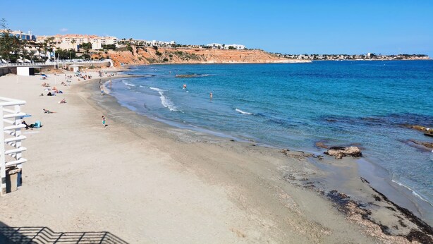 Une plage avec une eau d’un bleu limpide, un rivage sablonneux et des bâtiments en arrière-plan.