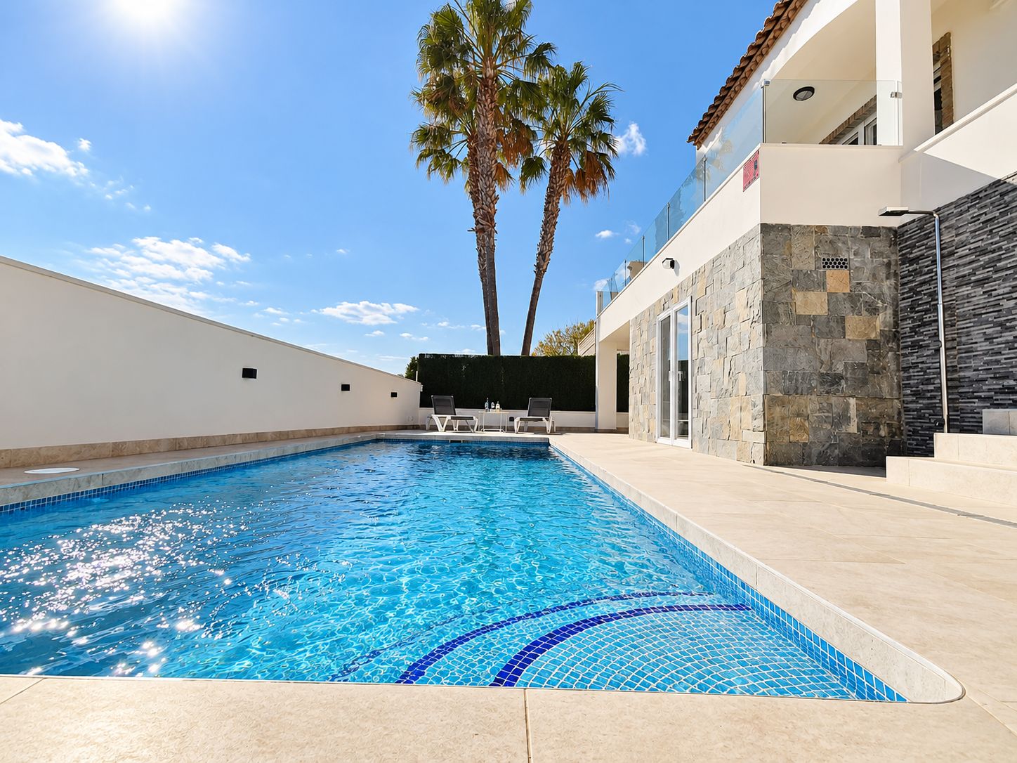 A swimming pool with clear blue water, surrounded by a white wall and a stone wall. There are two palm trees and a building with a tiled roof in the background.