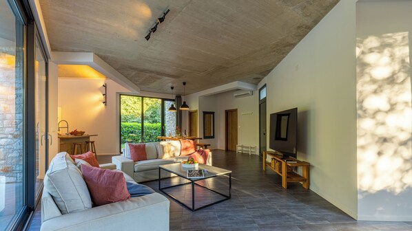 A spacious living room with a large sofa, a coffee table, and a television. There is a kitchen area with a dining table and chairs visible through an open door. - Unique Design Villa Doris (Umag)