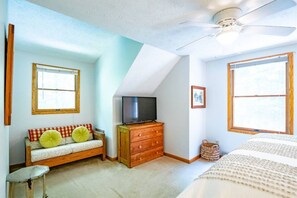 A bedroom with a sloped ceiling, a television on a wooden dresser, a sofa with cushions, and a window with a view. - Harvest Moon - Lake Access, Wooded Lot, Hot Tub! (Davis)
