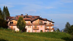 Un edificio de varios pisos con balcones, ubicado en un área verde con árboles y montañas al fondo.