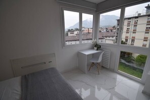 Une chambre à coucher comprenant un lit, un bureau, une chaise et de grandes fenêtres donnant sur des immeubles et des montagnes.