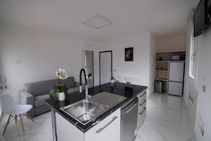 Une cuisine moderne dotée d’un îlot central, d’un lave-vaisselle encastré et d’un canapé gris dans le salon attenant.