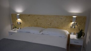 Un dormitorio con cabecera de madera, dos mesitas de noche con lámparas, una cama con ropa de cama blanca y una pequeña mesita con una planta.