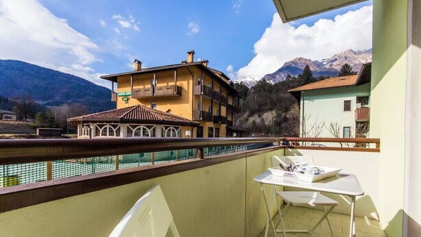 A balcony with a table and chairs, overlooking a swimming pool and buildings with balconies, set against a mountainous backdrop. - Family-Friendly Apartment Near Terme Comano (Ponte Arche)