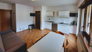 Une cuisine compacte avec des meubles blancs, un four à micro-ondes encastré et un lave-vaisselle. On y trouve une table à manger avec des chaises, un canapé et un radiateur.