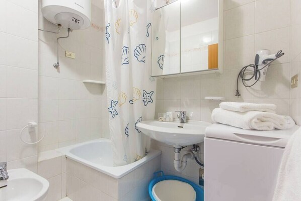 Un baño con bañera, lavamanos, inodoro y cortina de ducha con estampado de conchas marinas.
