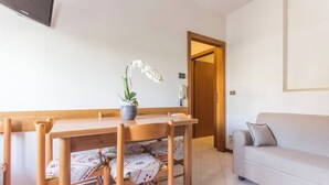 A dining area with a wooden table and chairs, a potted orchid, a sofa, and a door leading to another room.
