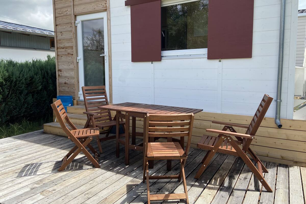 Una terraza de madera con mesa y sillas, una ventana con contraventanas marrones y una pared exterior blanca.