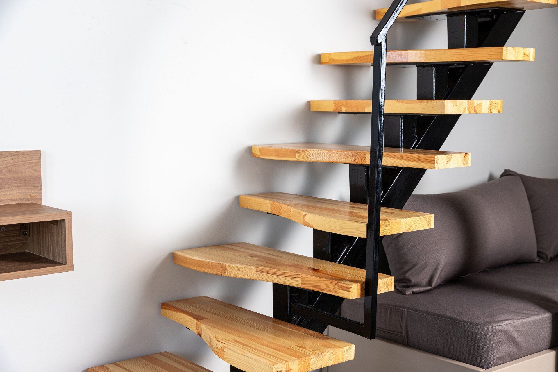 Una escalera moderna con peldaños de madera y barandilla de metal negro, que conduce a un sofá de color oscuro frente a una pared blanca.