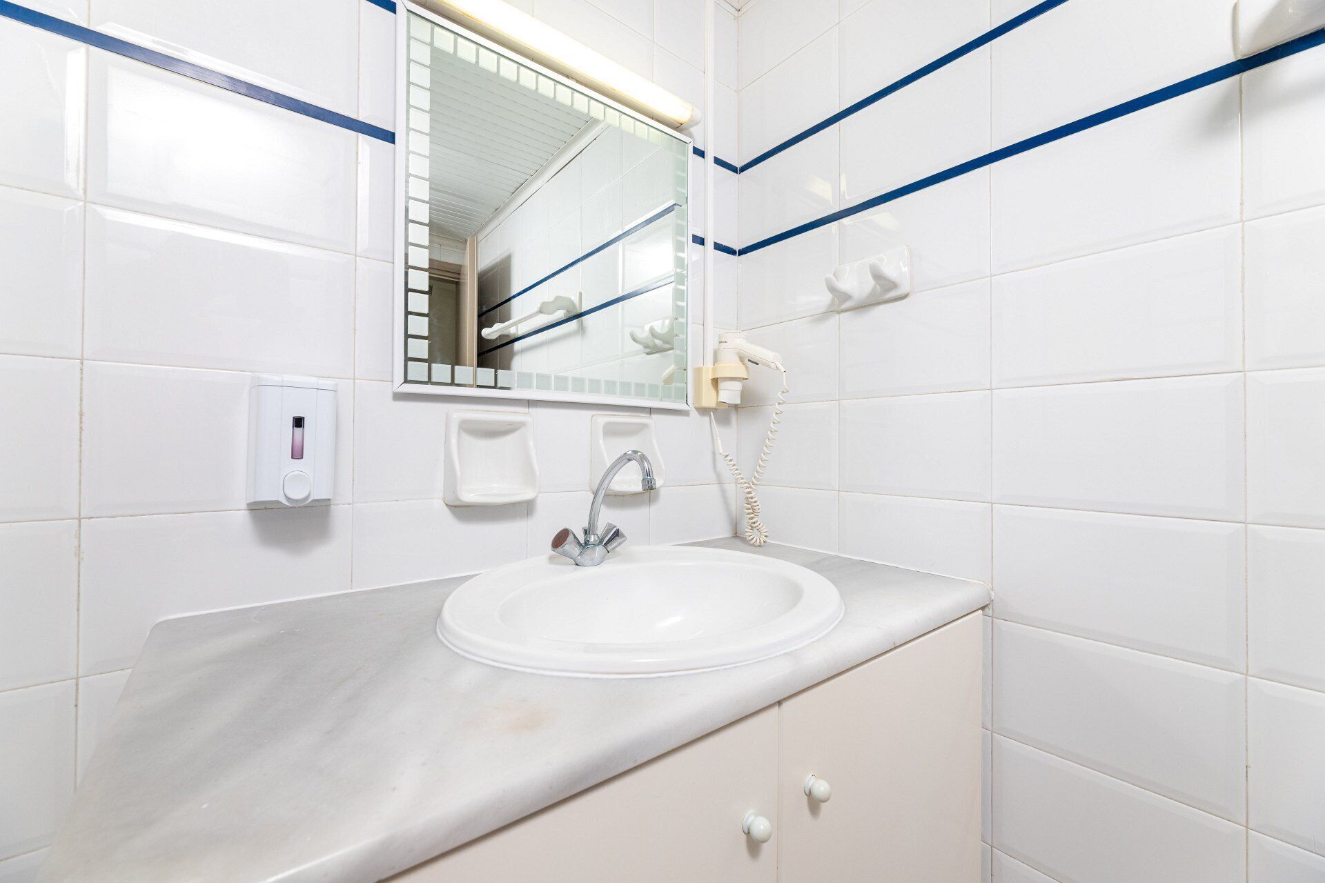 A bathroom with white tiles, a sink, a mirror, a hand dryer, and a soap dispenser.