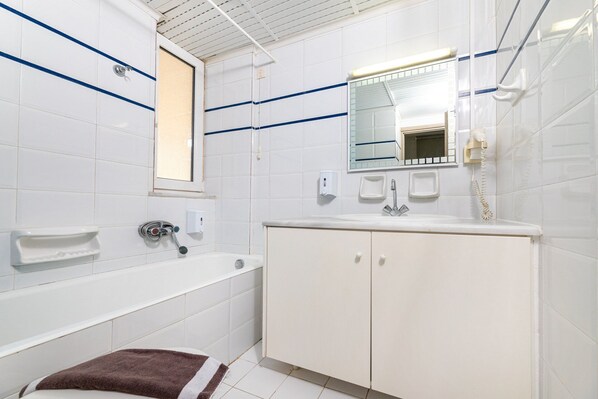 Un baño moderno con azulejos blancos, una bañera, un lavamanos con espejo y un teléfono.