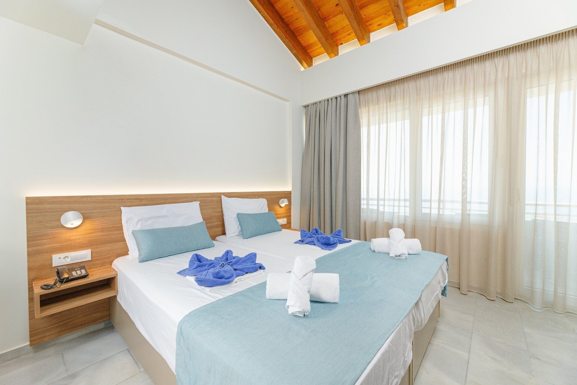 A hotel room with a large bed, a wooden headboard, a bedside table with a phone, and a window with curtains.