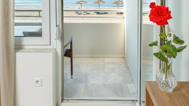 Habitación con puerta de vidrio corredera que da a un balcón con vista a la playa, una rosa roja en un florero y una mesita.