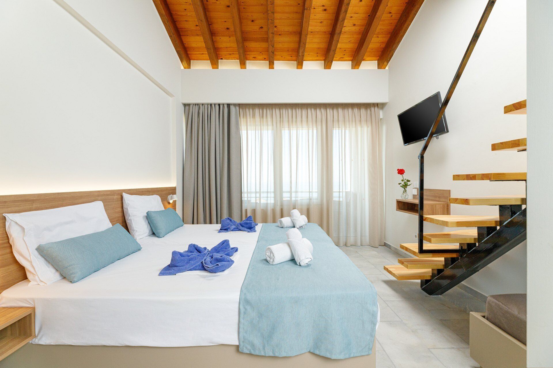 A hotel room with a bed, a TV, a staircase, and a window with curtains.