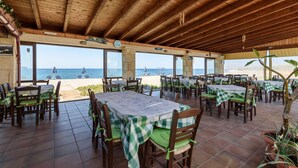 Un área de comedor al aire libre con mesas y sillas de madera, grandes ventanales con vistas a la playa y una vista del océano.