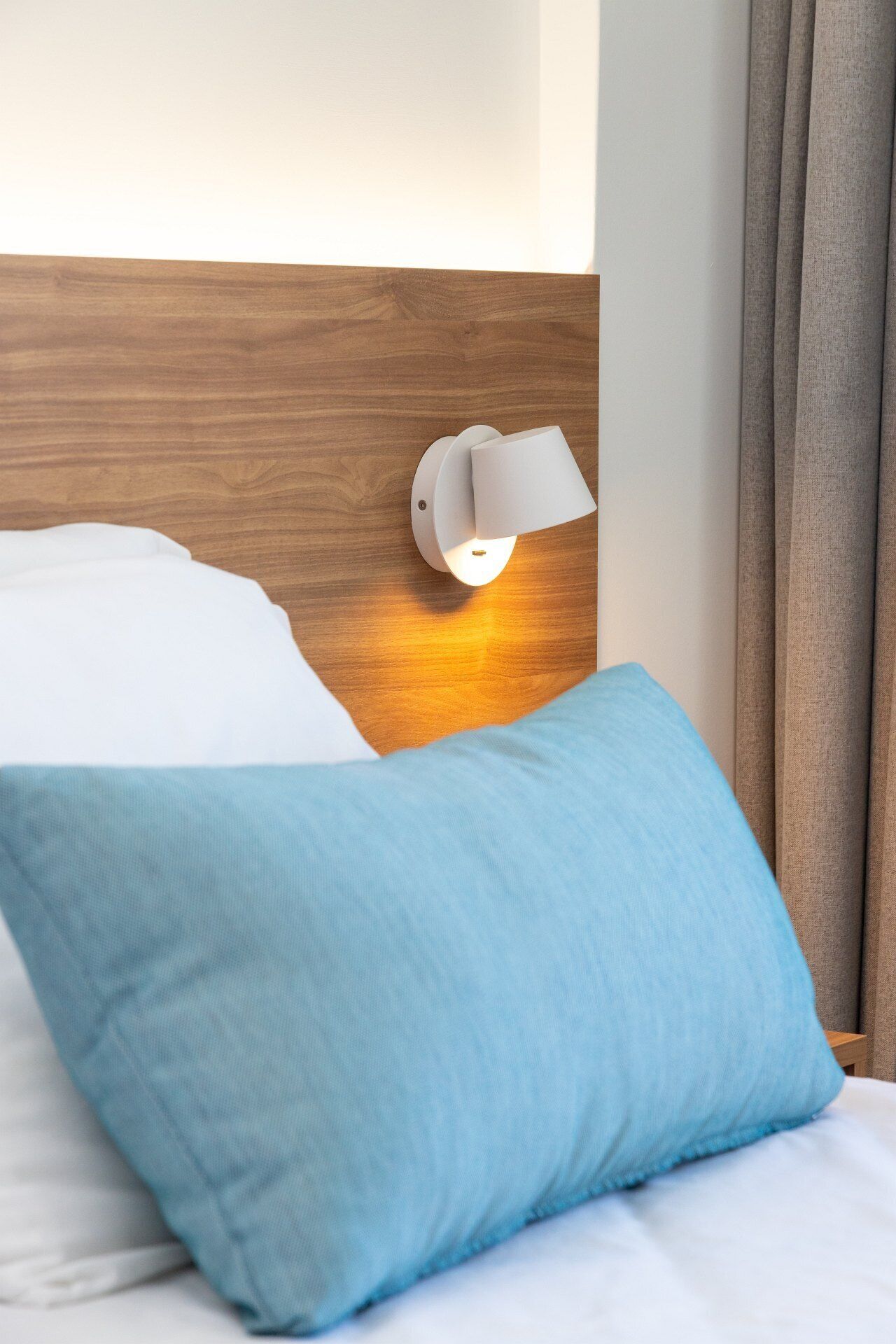 Una cama con un almohadón blanco y otro azul, un cabecero de madera y una lámpara fijada a la pared.