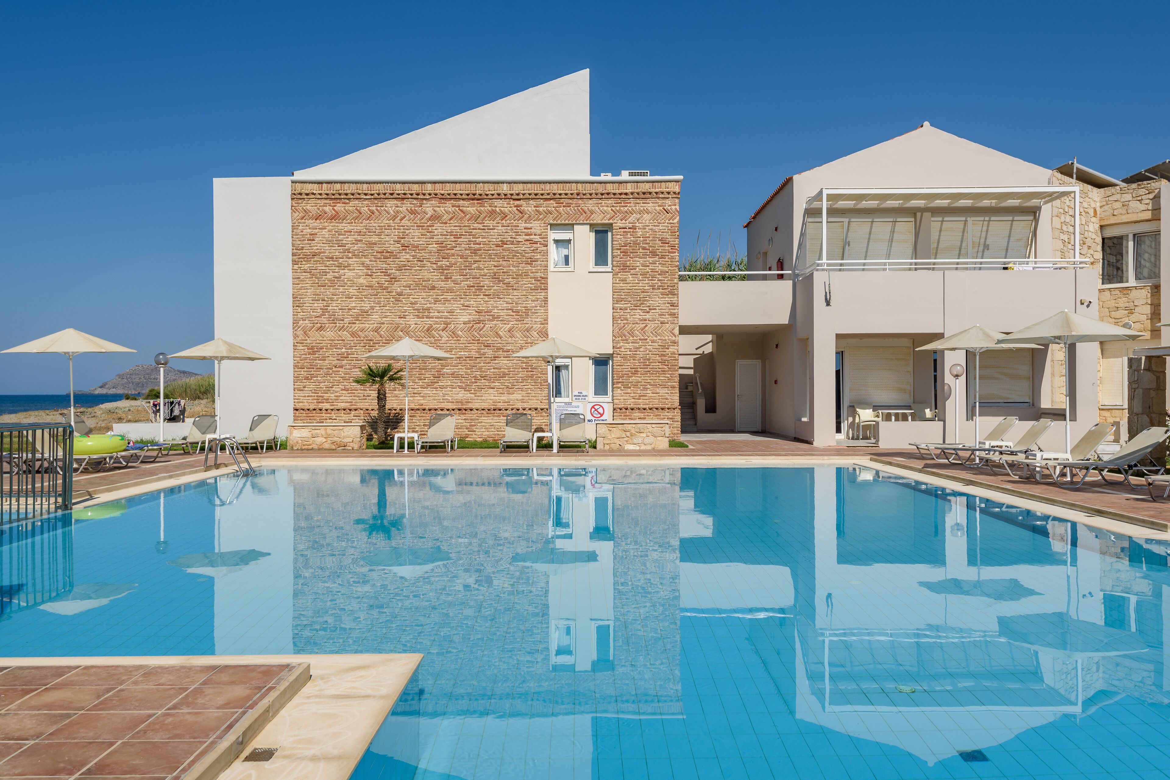 Uma piscina com espreguiçadeiras e guarda-sóis, um edifício com fachada de tijolo e um céu azul límpido.