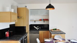 Kompakti keittiö, jossa on puiset kaapit, astianpesukone, musta työtaso sekä ruokailutila, jossa on lasipöytä ja violetit alustat.