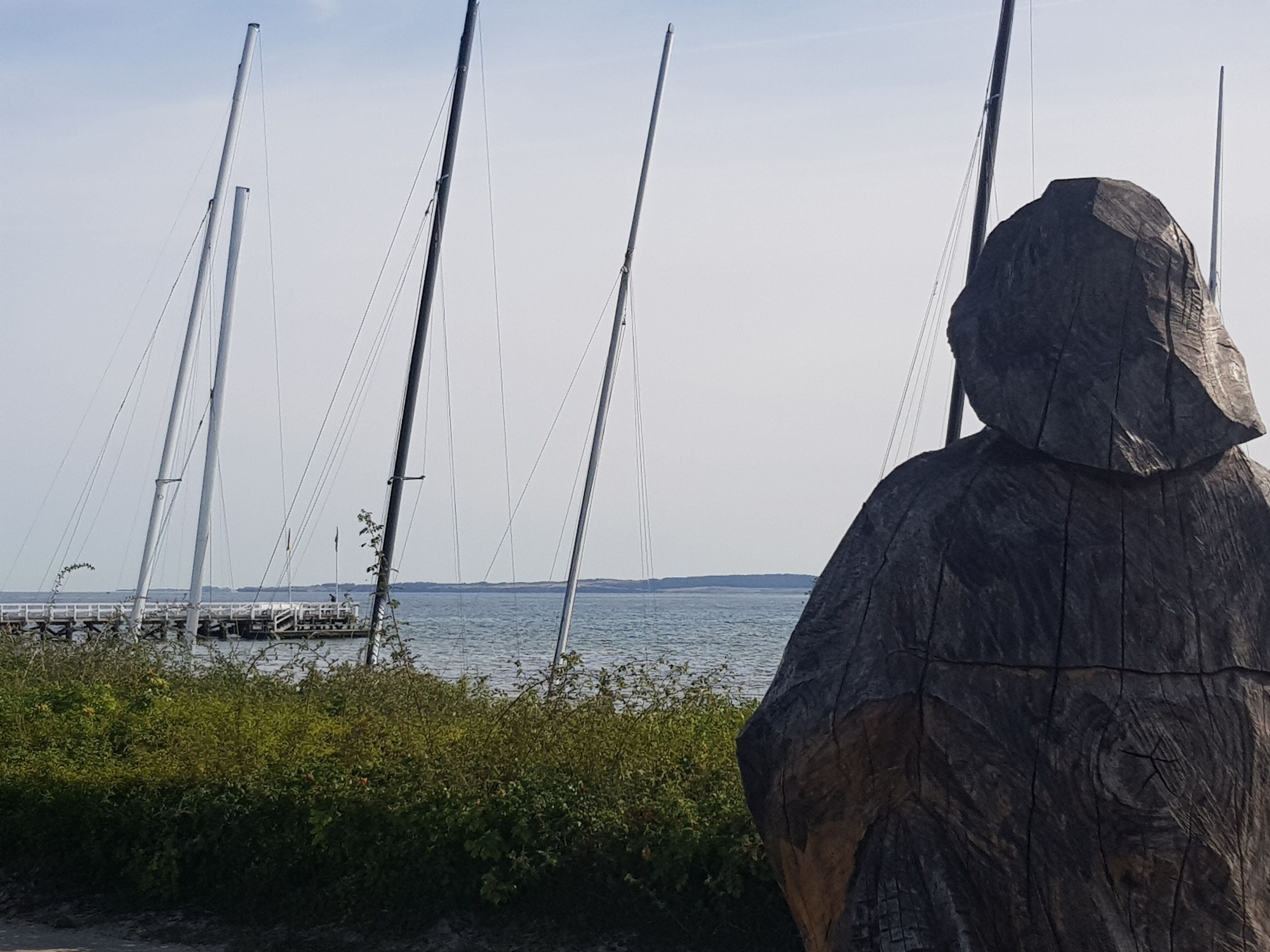 A sculpture of a figure with a hood, overlooking a body of water with sailboats.