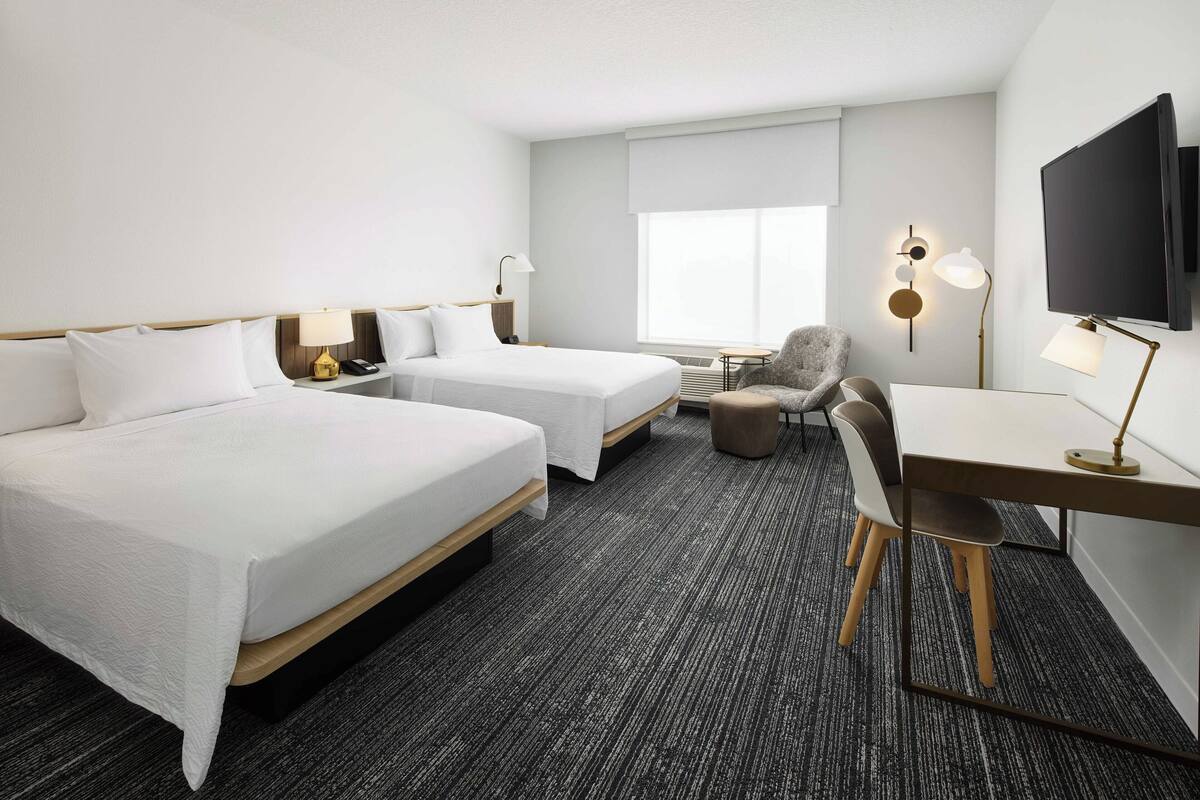 A hotel room with two beds, a desk, a chair, a TV, and a window.