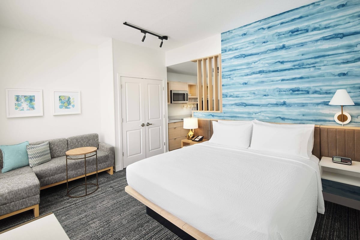 A hotel room with a large bed, a sofa, a small table, a desk, a lamp, a clock, and a view of the kitchen.