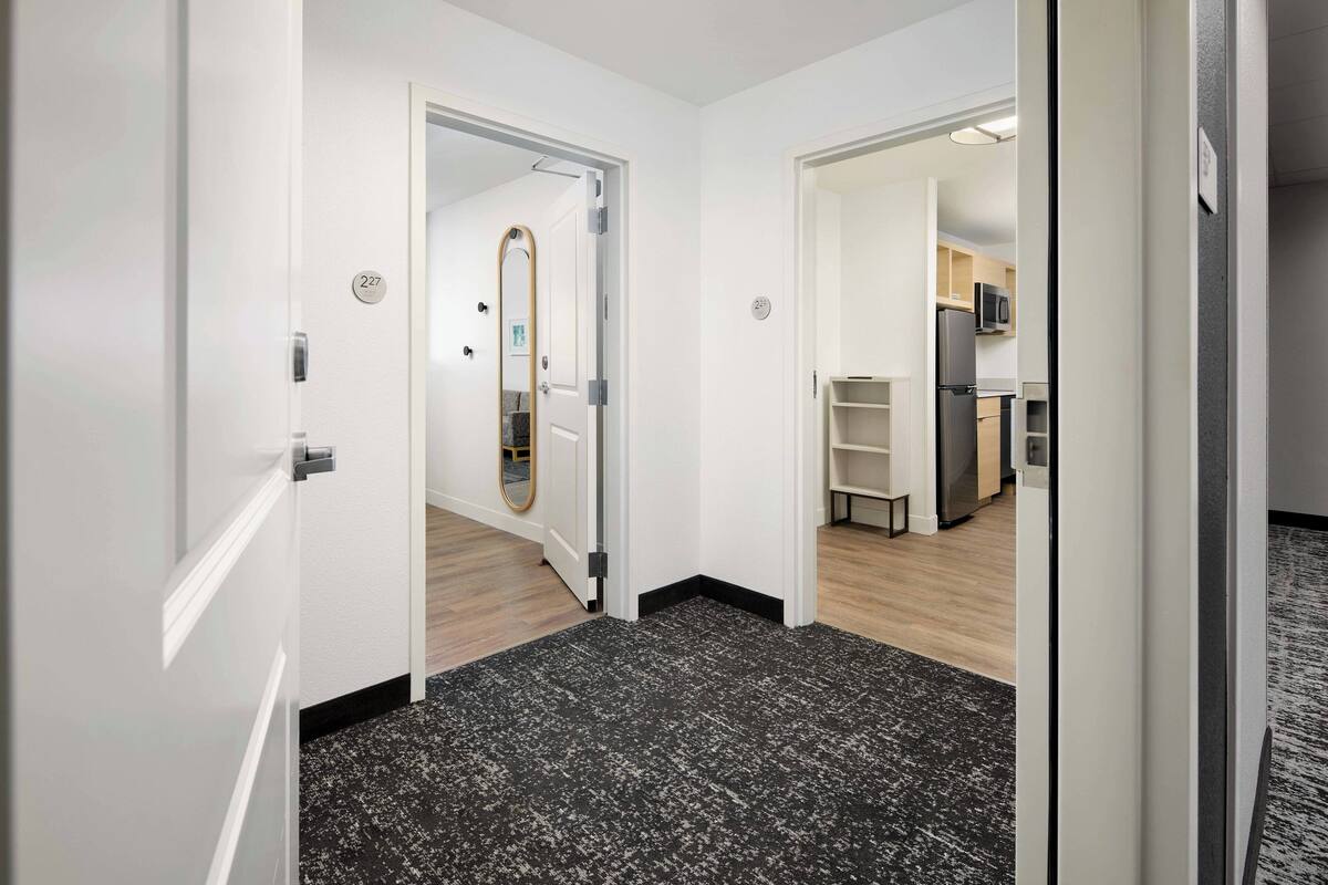 A hallway with a black and white patterned carpet, white walls, and a kitchen area visible through an open door.