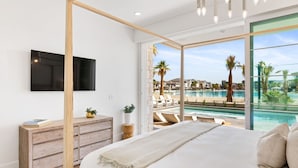 Un dormitorio con una cama grande, un televisor montado en la pared, un aparador de madera y vistas a una piscina y palmeras desde el exterior.
