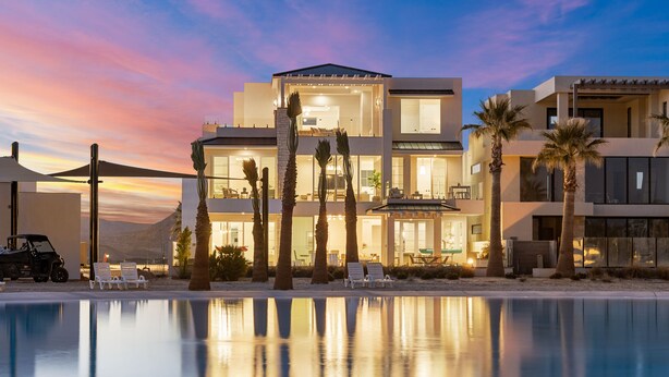 Un hotel moderno de varios pisos, con piscina, palmeras y un cielo al atardecer.