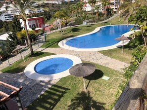 A swimming pool area with two pools, surrounded by grass and palm trees, with a view of residential buildings in the background. - BRIGHT PENTHOUSE SEA AND GOLF VIEWS TERRACE WIFI BARBECUE NEAR BEACH AND MARBELLA (Mijas Costa)