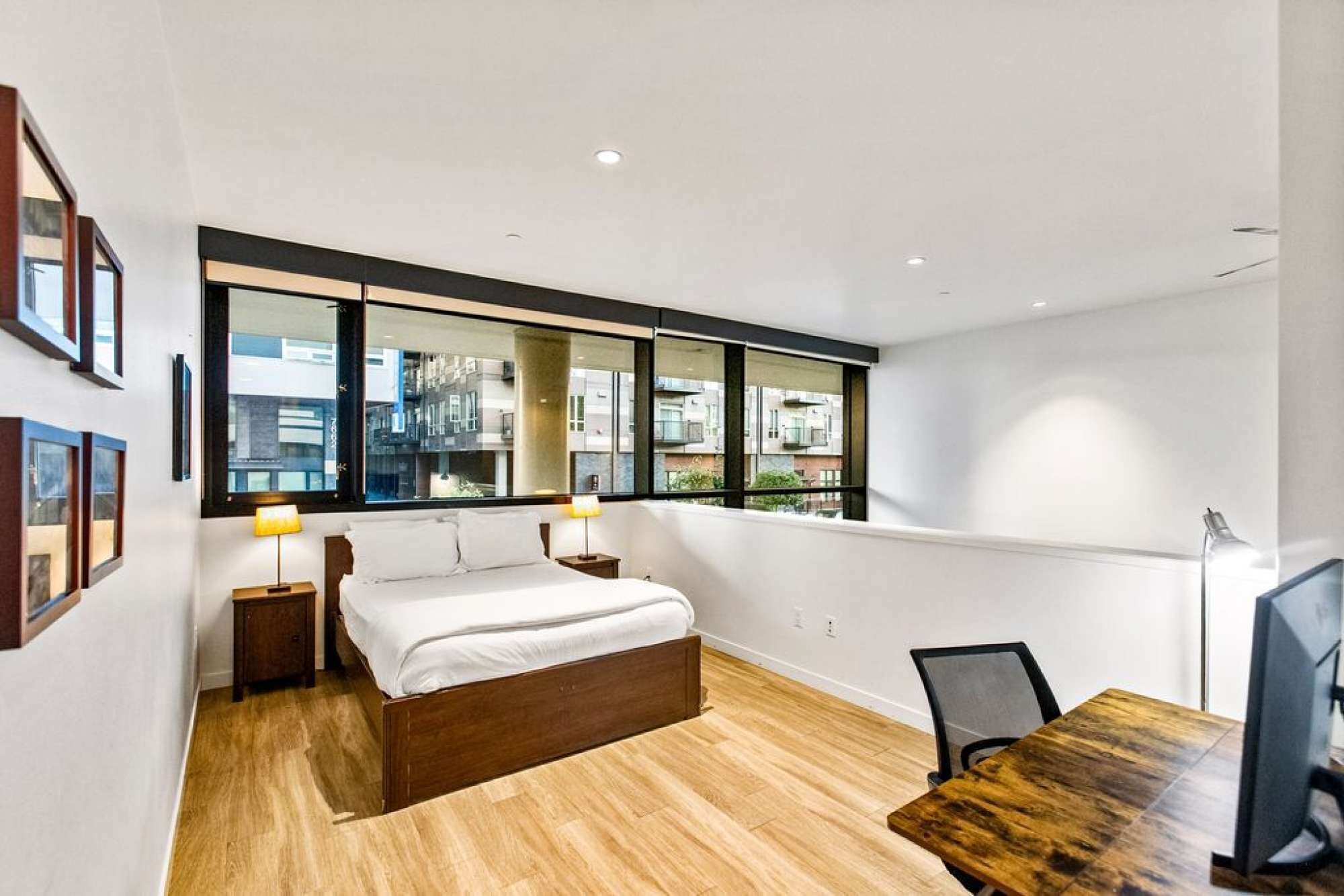Un dormitorio moderno con una cama grande, un escritorio y una silla. Hay dos mesitas de noche con lámparas, un ventanal con vistas a edificios y un televisor de pantalla plana sobre una mesa de madera.