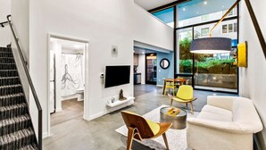 Una sala de estar moderna con una escalera, un televisor de pantalla plana, una zona de comedor con sillas amarillas y un ventanal que ofrece vistas al verdor.