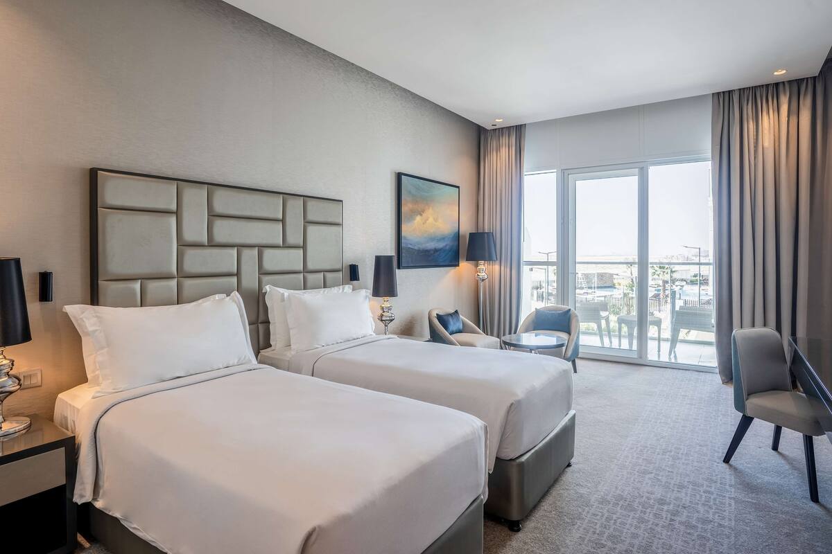 A hotel room with two beds, a seating area, a large window, and a painting on the wall.