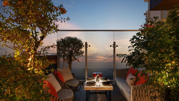 A terrace with a table set for dining, surrounded by wicker furniture and a view of the sunset over the water. - Radisson Dubai Damac Hills (Dubai)