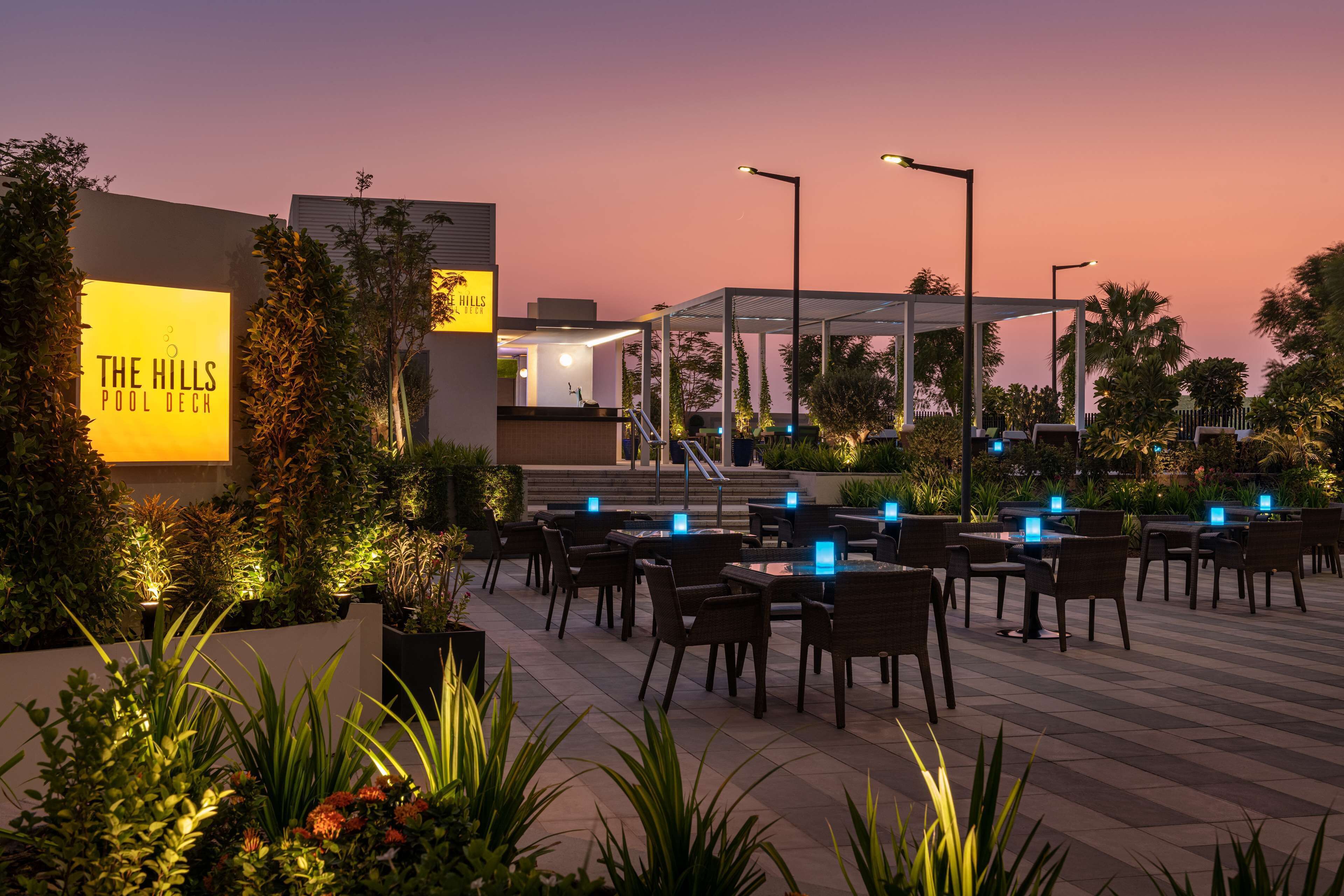 An outdoor seating area with tables and chairs, a sign for "The Hills Pool Deck," and a backdrop of trees and a sunset sky.